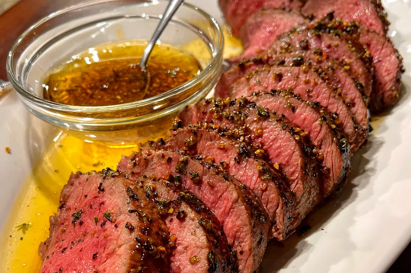 Premium ingredients for herb-crusted beef tenderloin including fresh beef, herbs, garlic, butter, and spices arranged on a wooden board